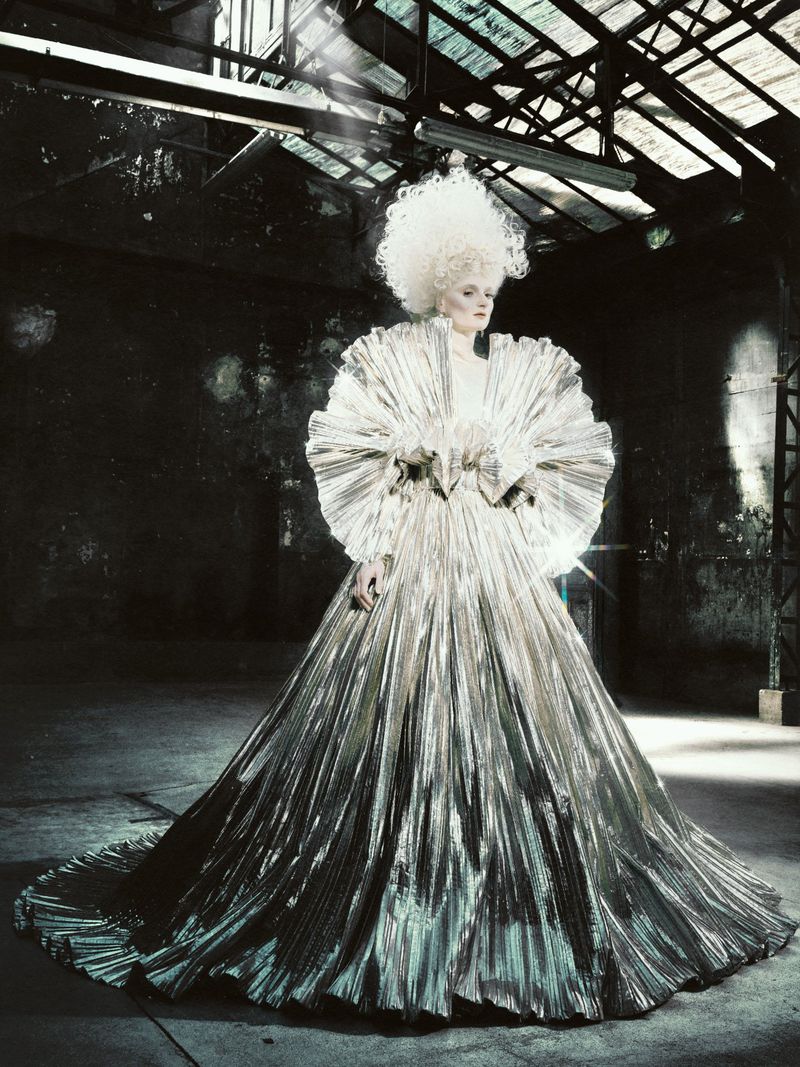 Model posing in an expansive pleated metallic gown with an oversized ruffled collar and white powdered hair in a warehouse.