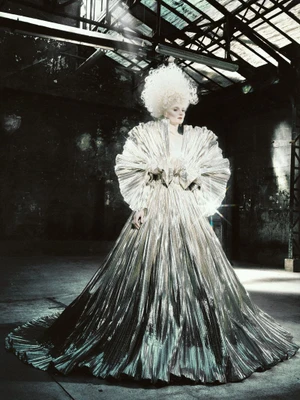 Model posing in an expansive pleated metallic gown with an oversized ruffled collar and white powdered hair in a warehouse.