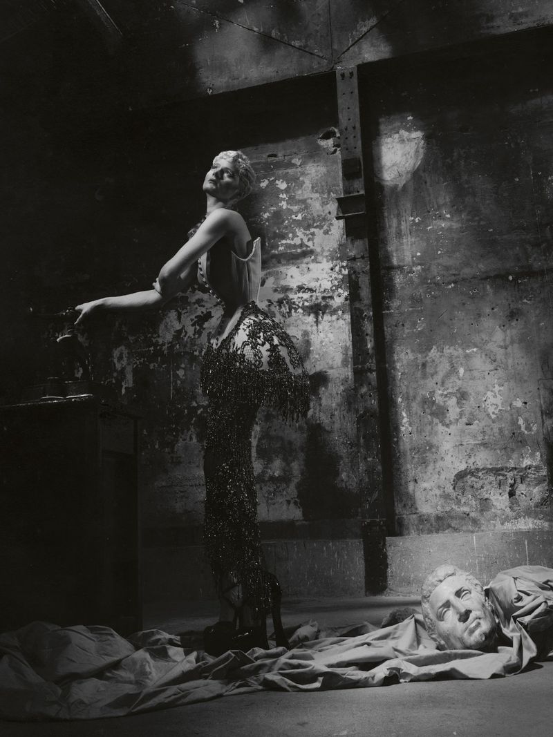 Monochrome fashion portrait of a model in a corset and beaded fringe gown posing in a warehouse setting beside a classical bust.
