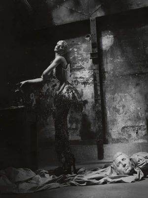 Monochrome fashion portrait of a model in a corset and beaded fringe gown posing in a warehouse setting beside a classical bust.
