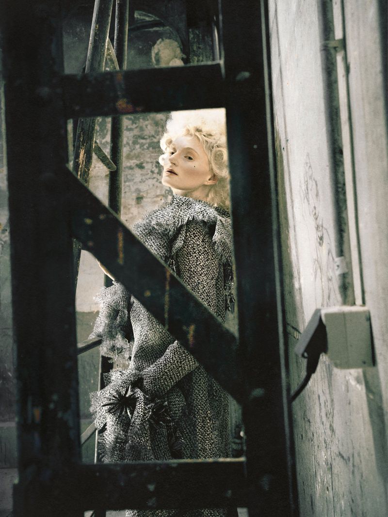 Model with styled blonde hair wearing a textured grey tweed outfit framed through a dark metal ladder frame in a rustic room.