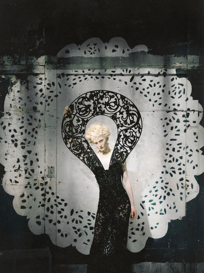 Model in a black lace dress and ornate graphic collar framed within a large decorative white paper doily cutout backdrop.