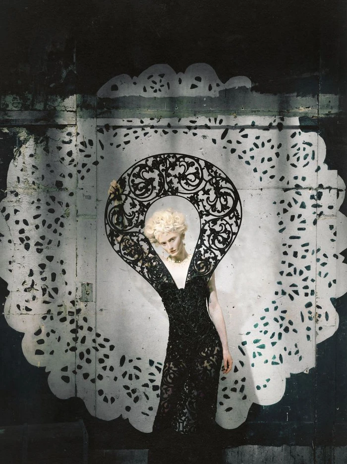 Model in a black lace dress and ornate graphic collar framed within a large decorative white paper doily cutout backdrop.
