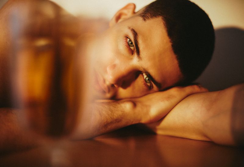 Close-up portrait of a young man resting his head on his arms under warm lighting with a blurred foreground.