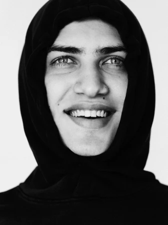 Black and white close up portrait of a smiling young man tightly framed in a black hood against a white background.