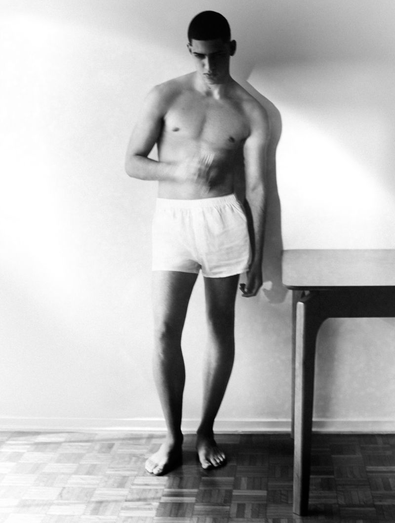 Monochrome portrait of a barefoot man in white boxer shorts standing by a table on a parquet floor.