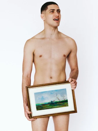 Shirtless man with short dark hair standing against a white backdrop holding a framed landscape painting covering his pelvis.