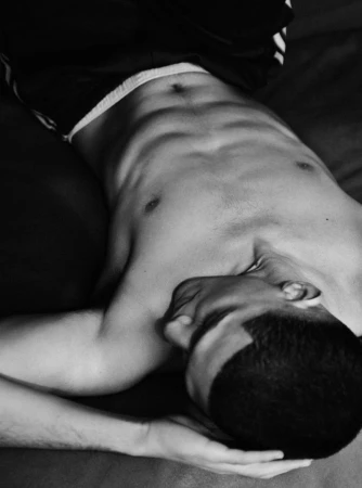 Black and white photograph of a shirtless man with a buzz cut lying down with his arm tucked behind his head.