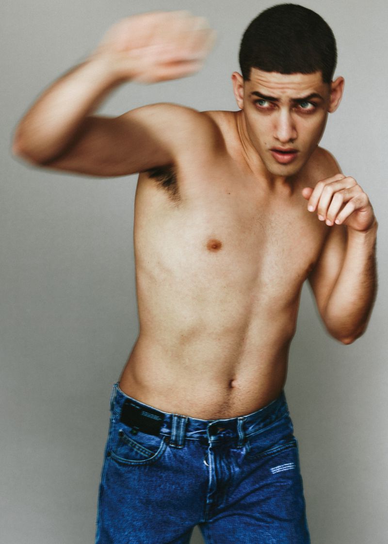 Shirtless man in blue denim jeans in a dynamic boxing punch stance against a plain grey studio backdrop.