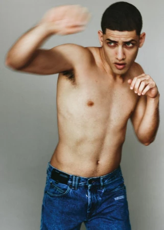 Shirtless man in blue denim jeans in a dynamic boxing punch stance against a plain grey studio backdrop.