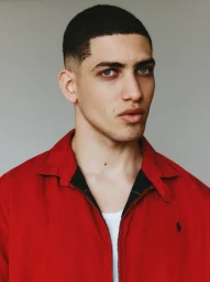 Portrait of a young man with a buzz cut wearing an open red zip-up jacket over a white undershirt against a grey studio backdrop.
