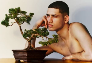 A shirtless young man with a buzz cut leaning thoughtfully beside a potted bonsai tree on a wooden surface against a plain backdrop.