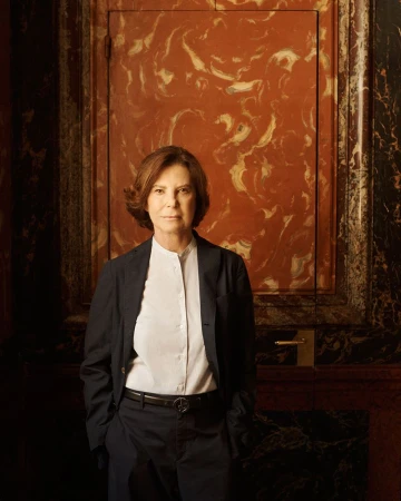 Silvana Armani wearing a tailored dark suit and white shirt, standing against a polished red marble wall.