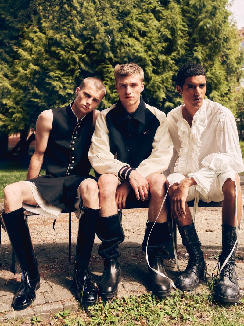 Three male models seated outdoors wearing styled black and white menswear including a ruffled shirt, varsity jacket, and tall boots.