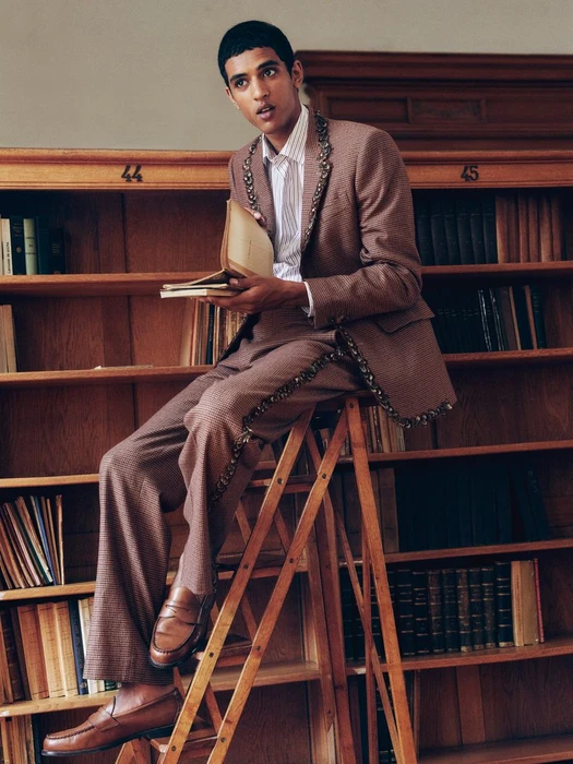 Model seated on a wooden library ladder wearing an embellished brown tailored suit and loafers while holding a book.