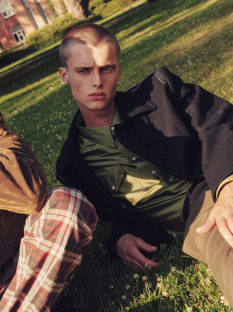 Model Elias Monstrey wearing an olive shirt and black jacket reclining on a sunny grassy lawn in an outdoor menswear editorial.