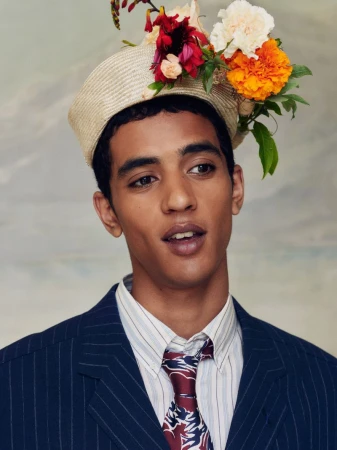 Man in a navy pinstripe suit and patterned tie wearing a straw hat adorned with colorful fresh flowers.
