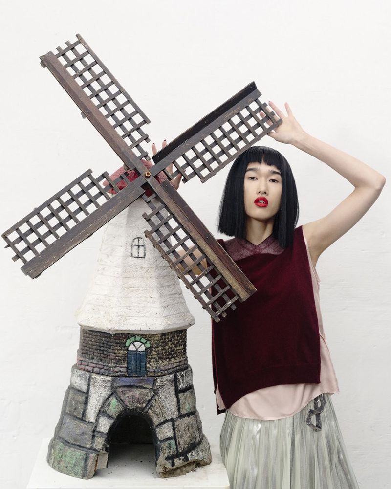 Model Asako Sato posing with a large windmill prop in a burgundy vest and pleated skirt against a white studio background.