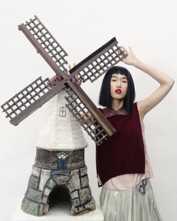 Model Asako Sato posing with a large windmill prop in a burgundy vest and pleated skirt against a white studio background.