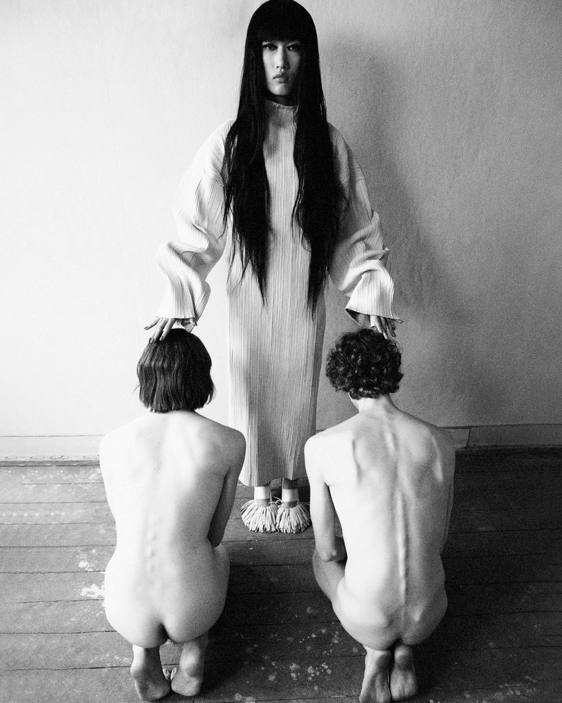 Model Asako Sato in a long white dress standing with hands on two kneeling figures in black and white