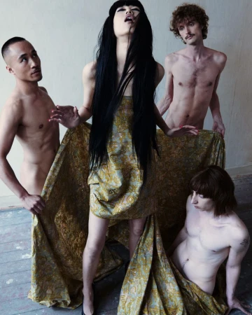 Model Asako Sato in a patterned gold brocade dress surrounded by three bare male models holding the draped fabric.