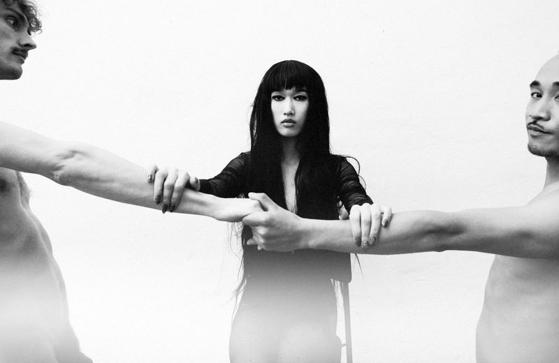 Model Asako Sato flanked by two shirtless men with interconnected arms in a black and white fashion editorial on a white backdrop.