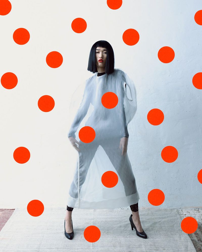 Model Asako Sato in a sheer translucent tunic and black pumps surrounded by bright red polka dots against a pale background.
