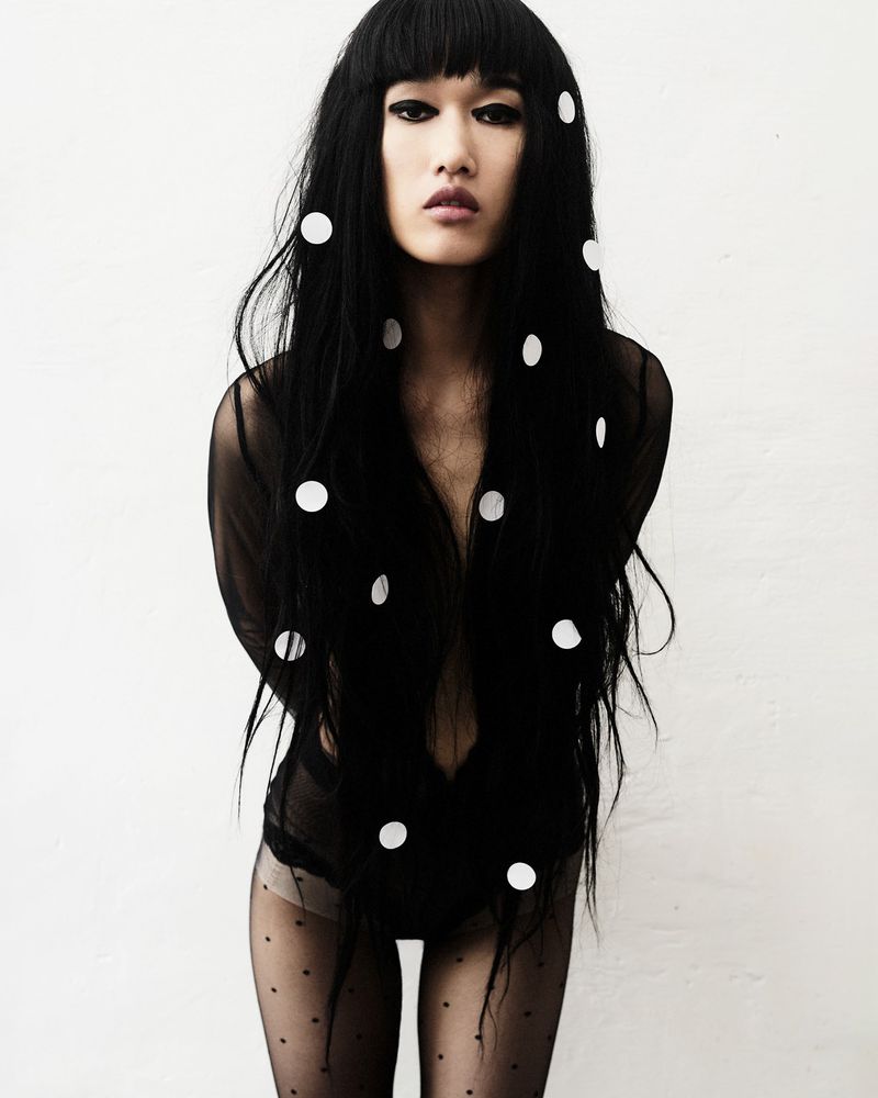 Model Asako Sato with long black hair adorned with white dots posing in sheer black bodysuit and tights against a white backdrop.