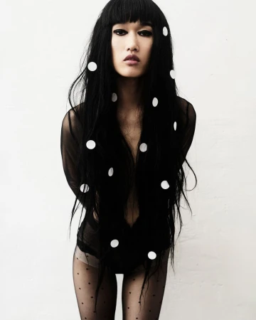 Model Asako Sato with long black hair adorned with white dots posing in sheer black bodysuit and tights against a white backdrop.