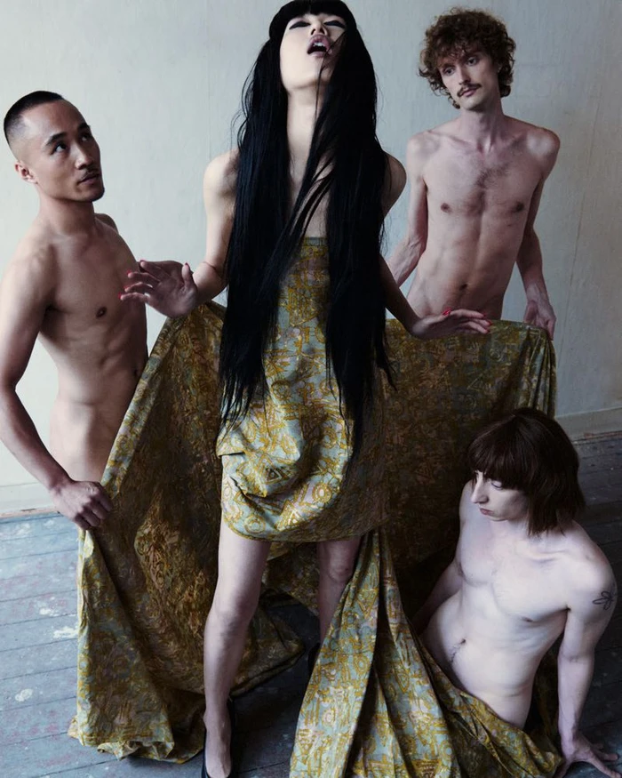 Model Asako Sato in a patterned gold drape surrounded by three shirtless male models in an editorial scene.