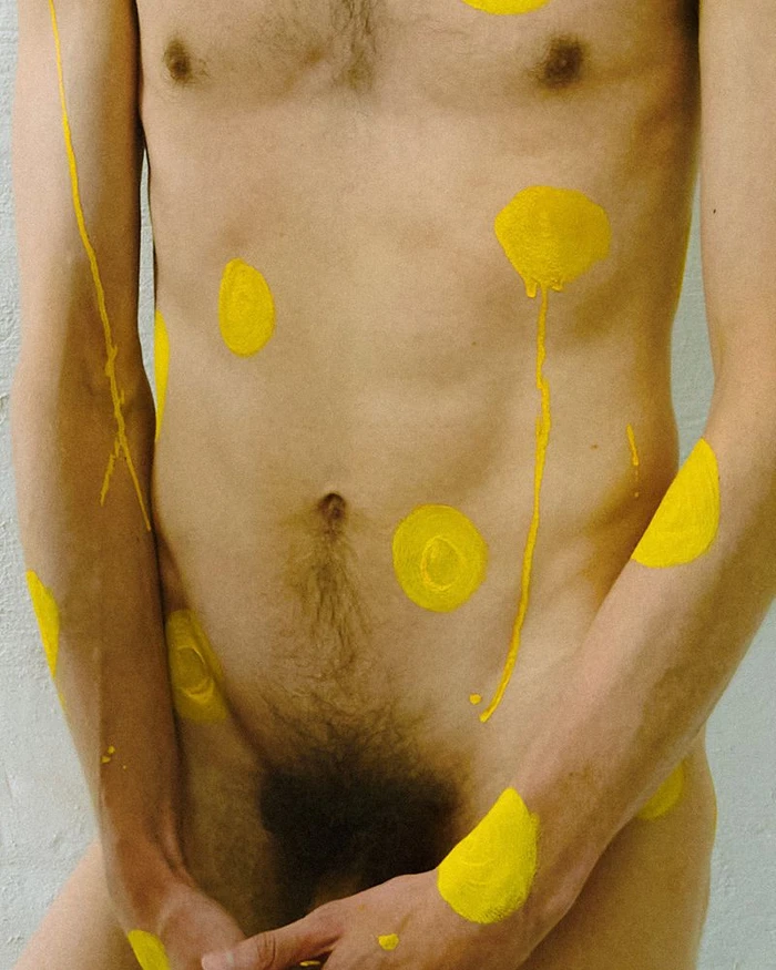 Close-up of a nude male torso and arms decorated with painted bright yellow circles and dripping paint marks.