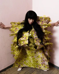 Model Asako Sato in a voluminous green polka-dot sculptural gown standing with arms outstretched in a pink corner.