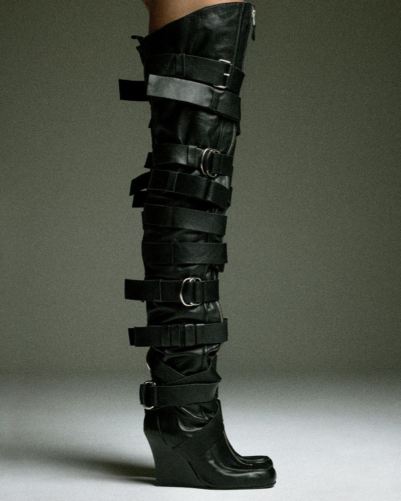 Close-up of a model wearing a tall black leather thigh-high boot with multiple buckled straps and a wedge heel against a studio backdrop.