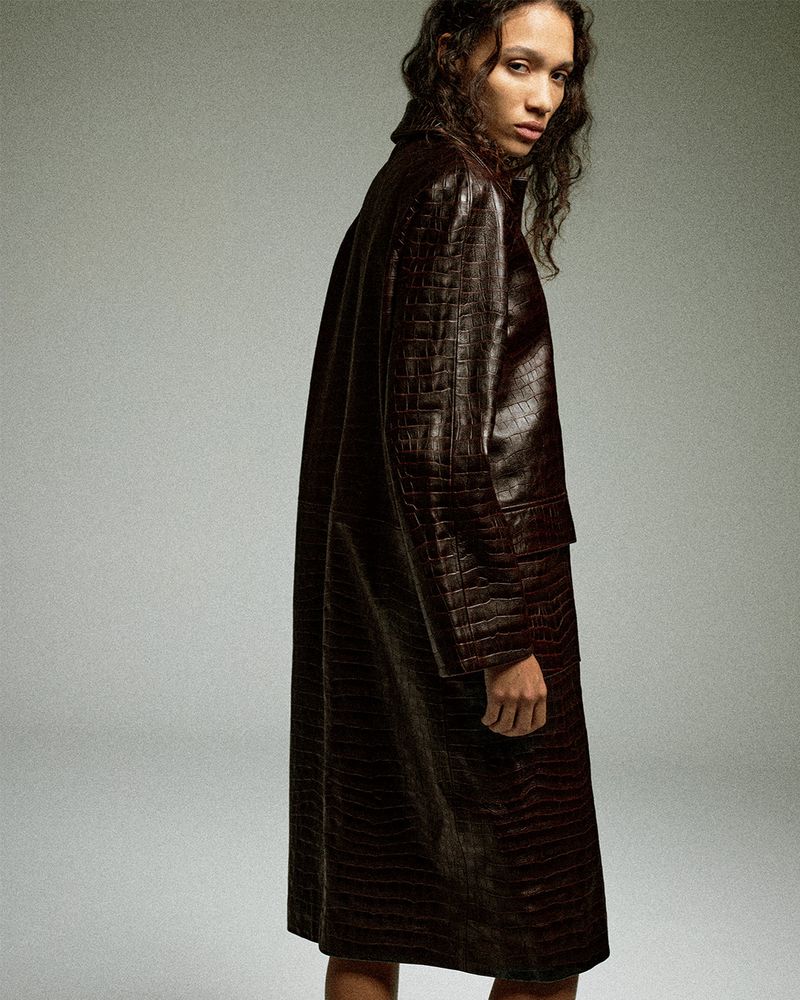 Model Mauria Caetano wearing a long dark brown crocodile-embossed leather coat against a seamless grey studio backdrop.