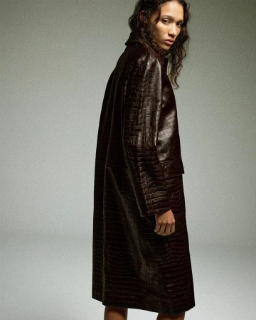 Model Mauria Caetano wearing a long dark brown crocodile-embossed leather coat against a seamless grey studio backdrop.