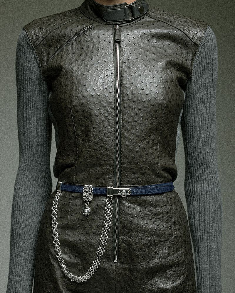 Cropped view of a model in a textured dark ostrich leather zip vest over grey ribbed knit sleeves with a belted chain.