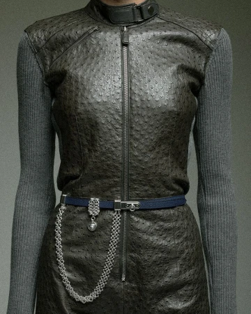 Cropped view of a model in a textured dark ostrich leather zip vest over grey ribbed knit sleeves with a belted chain.