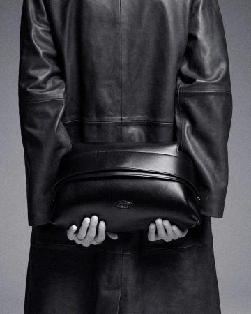 Model wearing an oversized black leather coat holding a black leather handbag behind her back against a studio backdrop.
