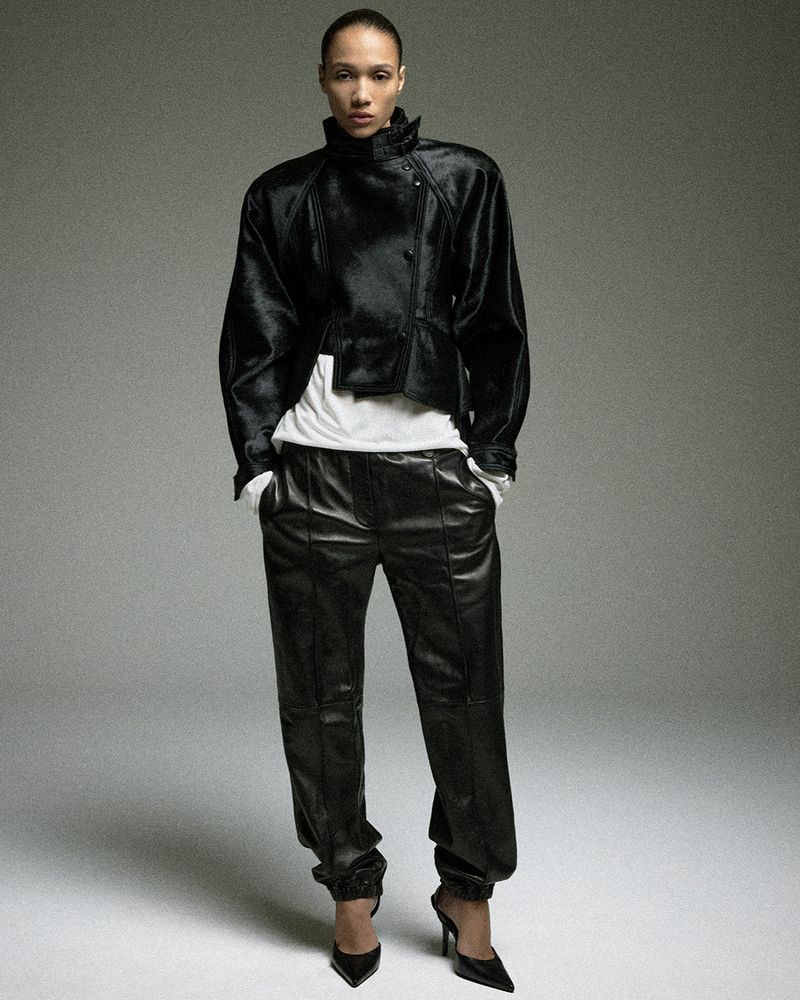 Model Mauria Caetano poses in a black leather jacket, matching loose leather trousers, and pointed stiletto heels against a grey backdrop.