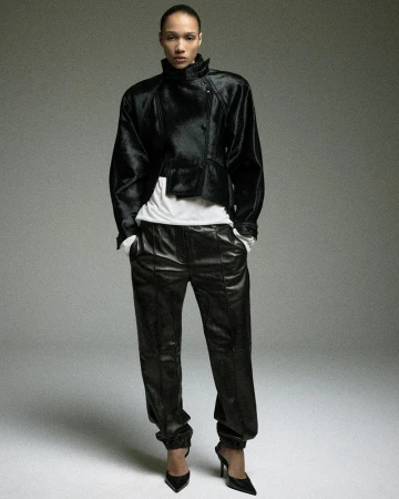 Model Mauria Caetano poses in a black leather jacket, matching loose leather trousers, and pointed stiletto heels against a grey backdrop.