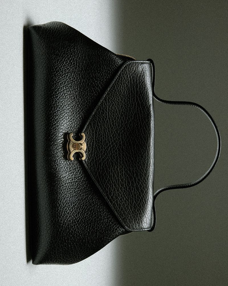 Black textured leather handbag with gold metallic clasp and curved top handle against a neutral grey background.