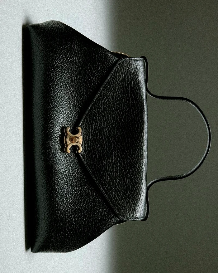 Black textured leather handbag with gold metallic clasp and curved top handle against a neutral grey background.