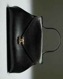 Black textured leather handbag with gold metallic clasp and curved top handle against a neutral grey background.