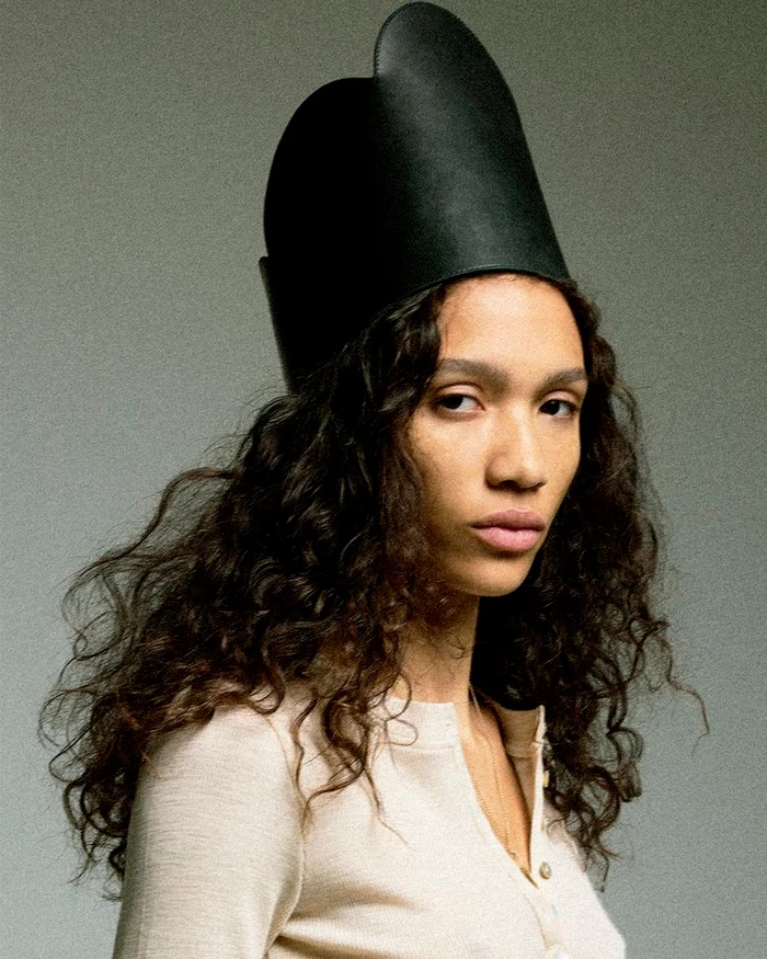 Mauria Caetano wearing a tall black leather headpiece and cream top against a grey seamless background.