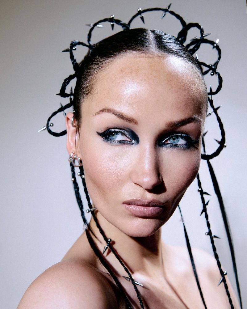 Close-up beauty portrait of a woman with an avant-garde spiked braided headpiece and bold black eyeliner against a pale background.