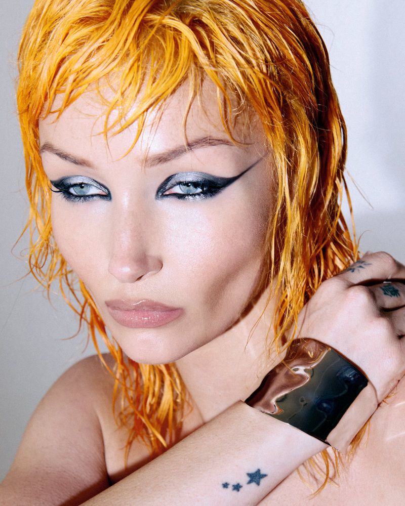 Close-up portrait of a woman with wet orange hair, bold winged eyeliner, and a metallic chrome cuff bracelet against a plain background.