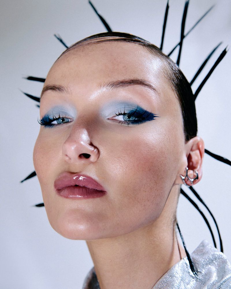 Close-up portrait of a woman with spiky black hair, blue winged eyeshadow, and a silver ear cuff against a plain studio background.