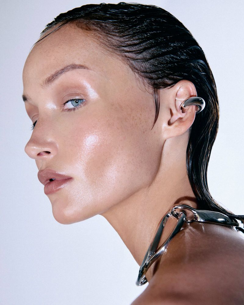 Close-up profile of a woman with slicked-back wet-look hair wearing a sculptural silver ear cuff and metal collar against a white background.