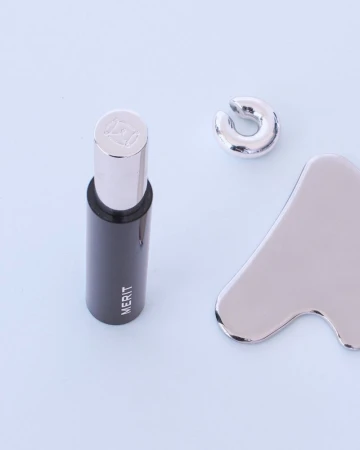 Merit cosmetic tube standing upright next to a silver metallic gua sha tool and silver ear cuff on a pale surface.