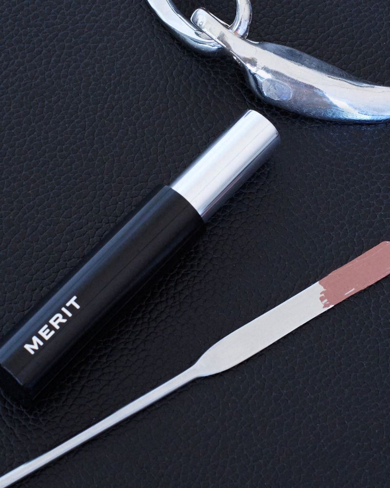 Merit makeup tube beside a silver cosmetic spatula with a nude product swatch on a black textured surface.